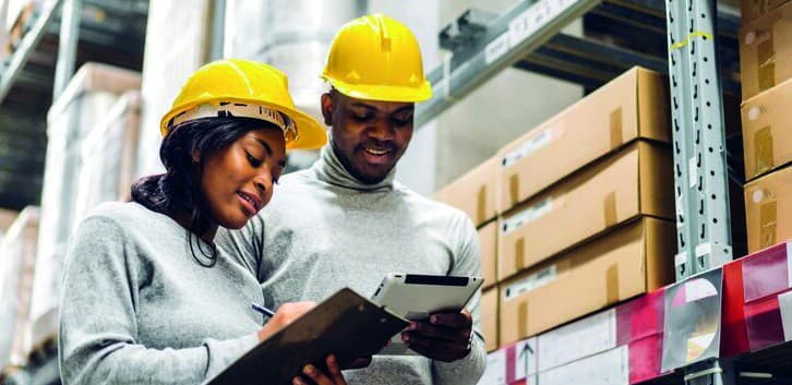 Two Gravan team members in hard hats reviewing stock on a tablet in an industrial warehouse aisle.